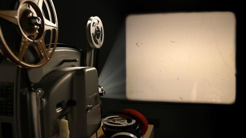 A vintage 8mm film projector projects a blank image with film dust and scratches onto a wall beside a stack of film reels