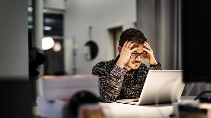 Young casual businessman wearing glasses is sitting in front of his notebook holding his head pondering over his work. Office equipment and another computer is in front of him.