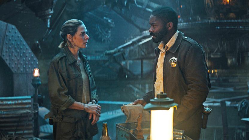 Rebecca Ferguson and David Oyelowo stand in a darkened industrial space with large steel girders and machines in this promo image for the Apple TV+ series 'Silo.'