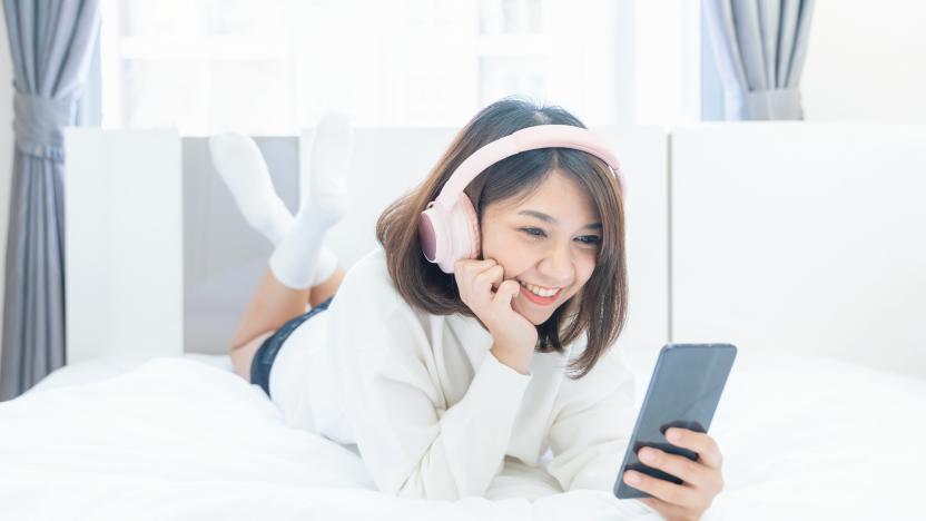 Beautiful young woman with headphones relaxing on the bed, Female listening to music using smart phone, Chill out and leisure concept