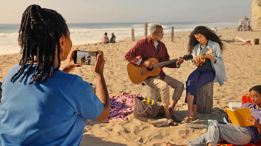 An over-the-shoulder view of a woman taking a spatial video with her iPhone of a man and woman playing guitar at the beach. 