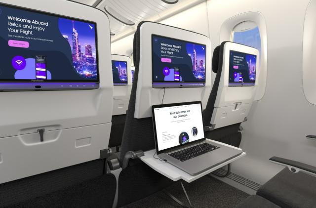 The backsides of plane seats in economy class installed with Panasonic Avionics' Astrova in-flight entertainment system screens. The screens display a notice that says "Welcome Aboard, relax and enjoy your stay." One tray is unfolded with an open laptop on top of it. 