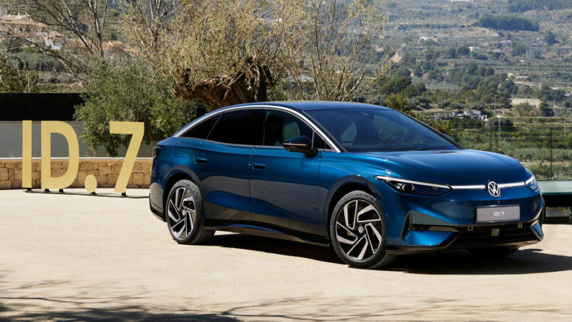 The Volkswagen ID.7 sedan (blue) sitting on a driveway with a sign reading "ID.7" sitting behind it. Trees and a valley visible behind both.