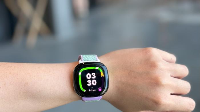 The Fitbit Ace LTE on a wrist held in mid-air, with the time 03:30 on the screen, surrounded by a green snake-like graphic.