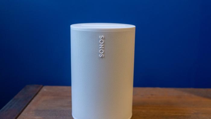 Photo of the Sonos Era 100 smart speaker (white) sitting on a wooden table in front of a purple wall.