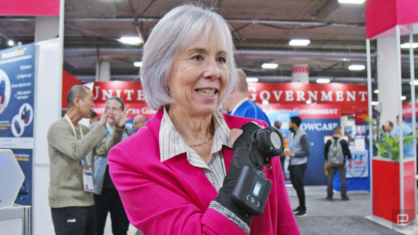 A person in a pink blazer wearing the GyroGlove hand stabilizer, holding their forearm and fist up to their chest. 