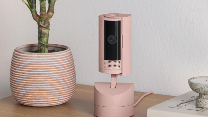 Image of the new Ring indoor pan-tilt camera on a movable base in "blush" which is a fancy word for pink. It is on a table next to a plant pot and some fancy coffee-table books about art that you can't read because there's a dish on top of it.