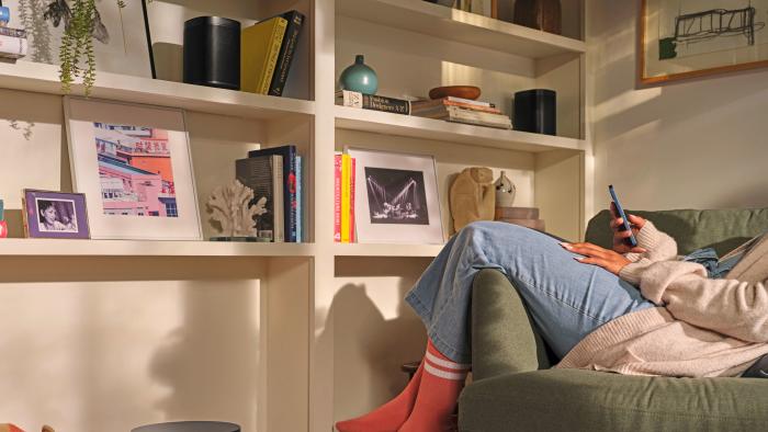 The view of someone's living room with a Sonos speaker on the book shelf and a sub on the floor next to a couch.