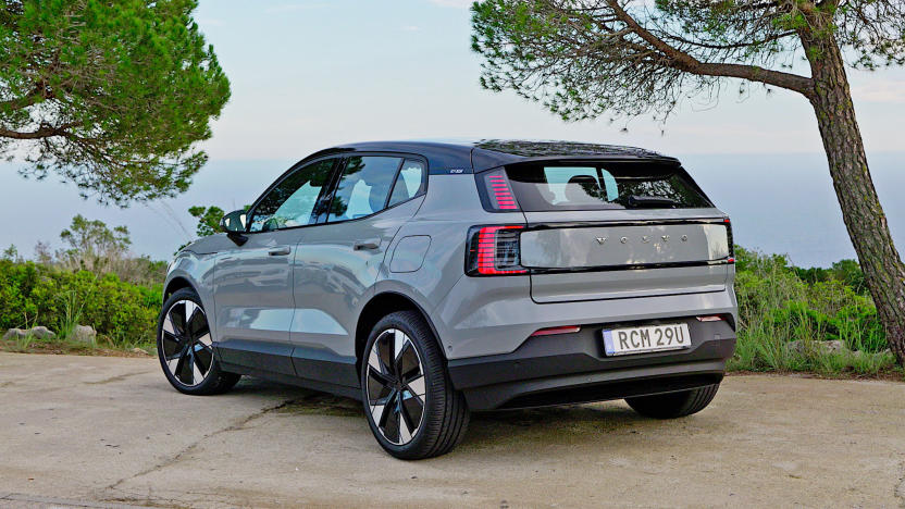 A grey Volvo EX30 seen from behind is parked in a cement lot next to a small foliage covered area overlooking a large expanse of water.