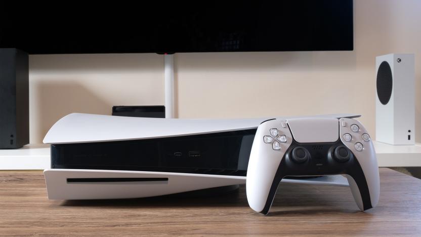 The white and black PlayStation 5 console is on a wooden coffee table with a TV and Xbox in the background. A PlayStation controller sits in front of the console. 