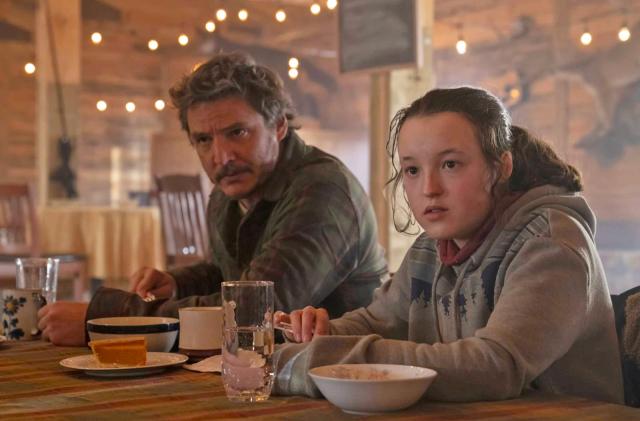 An image from the TV series 'The Last of Us' showing Ellie and Joel seated at a table eating breakfast from bowls, while looking at someone off camera.