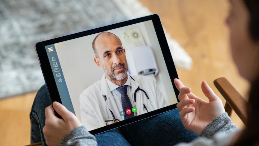 Back view of young woman making video call with her doctor while staying at home. Close up of patient sitting on armchair video conferencing with general practitioner on digital tablet. Sick girl in online consultation with a mature physician.